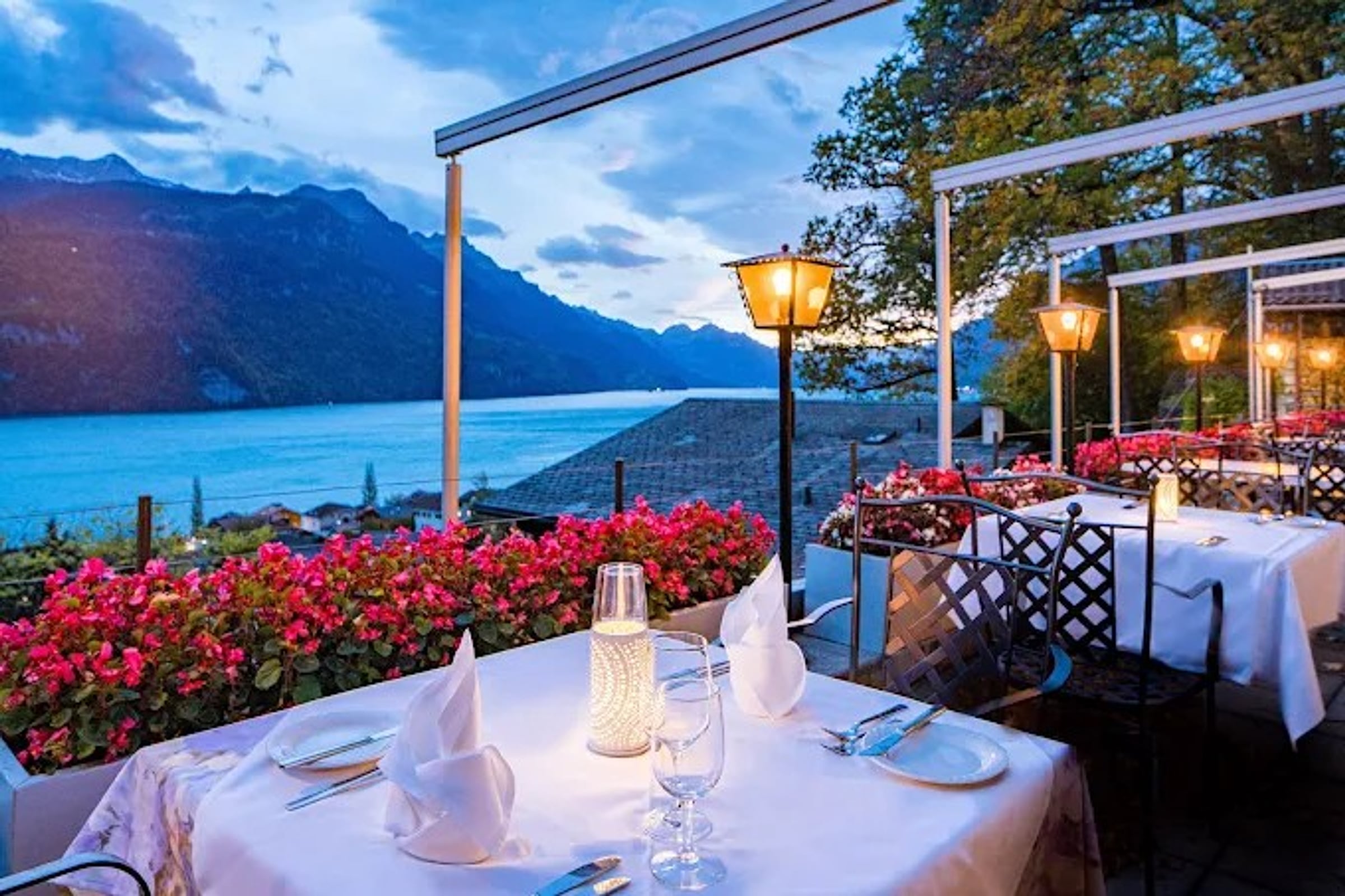 Die Terrasse von Restaurant Lindenblüte in der Abenddämmerung, mit leuchtenden Laternen, einem elegant gedeckten Tisch, leuchtend roten Blumen und der Bergsilhouette über dem Brienzersee